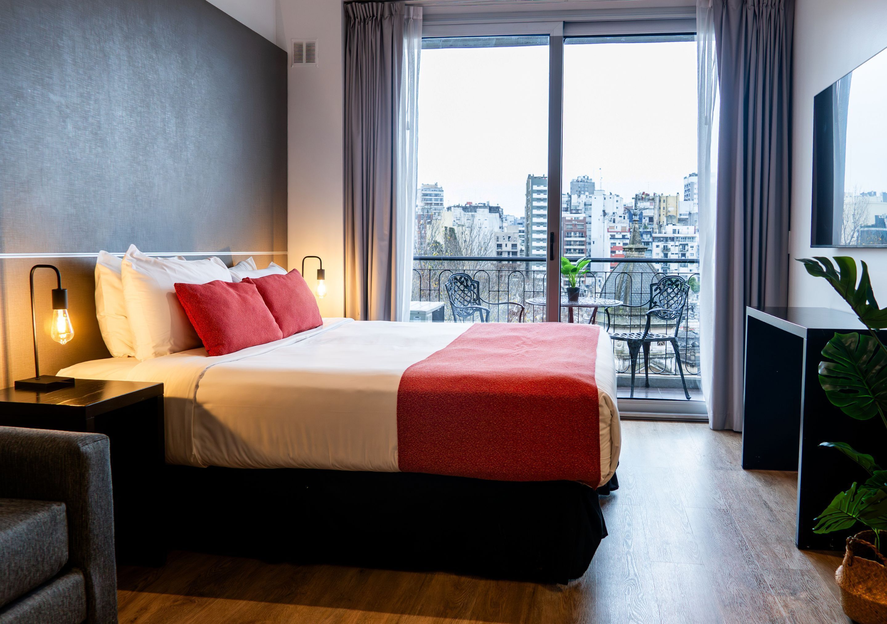 A hotel room with a large bed, a balcony with a city view, a desk with a lamp, and a chair.