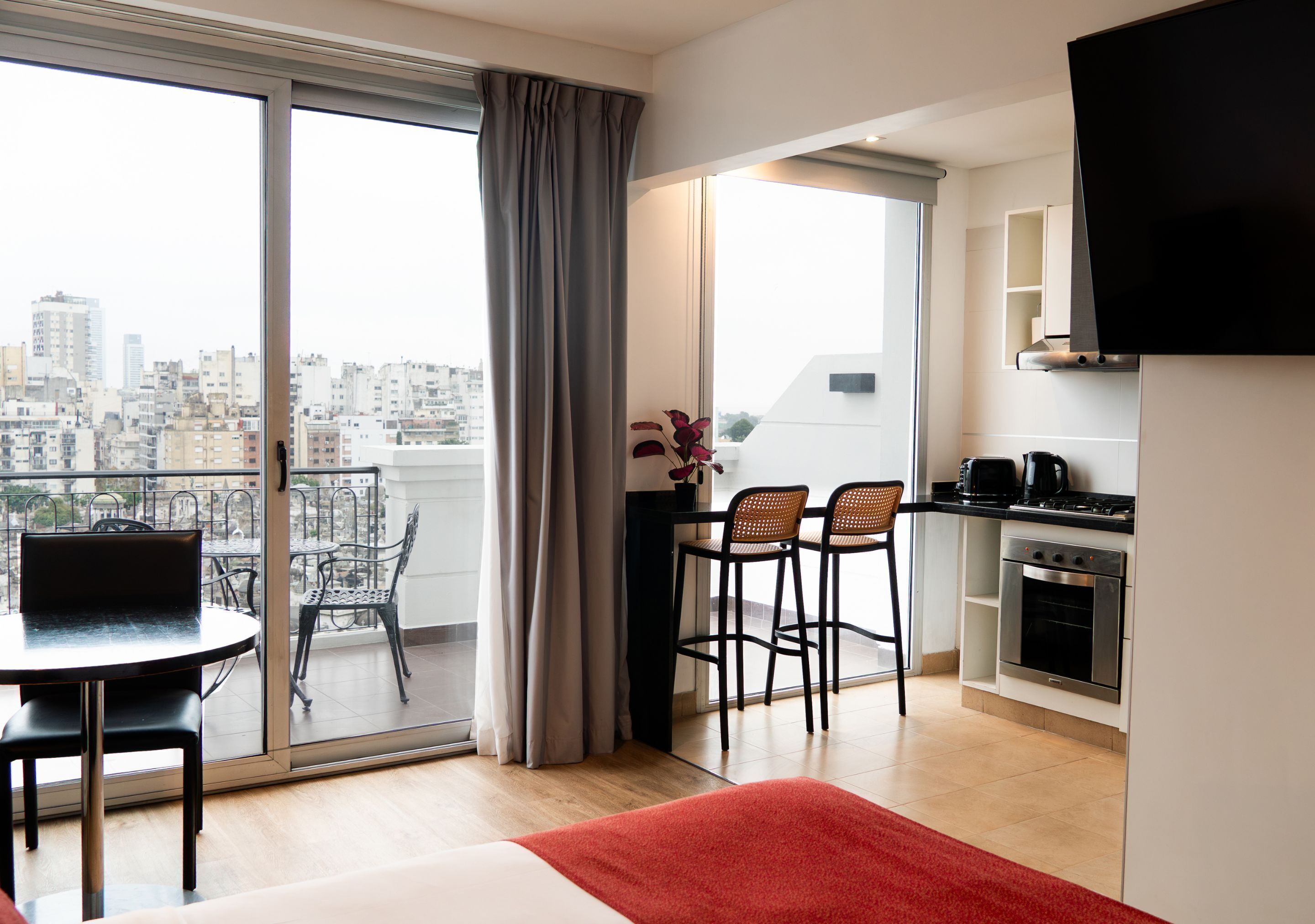A modern apartment with a balcony, a dining area with chairs, a kitchen with a stove, and a TV mounted on the wall.