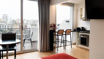 A modern apartment with a balcony, a dining area with chairs, a kitchen with a stove, and a TV mounted on the wall.
