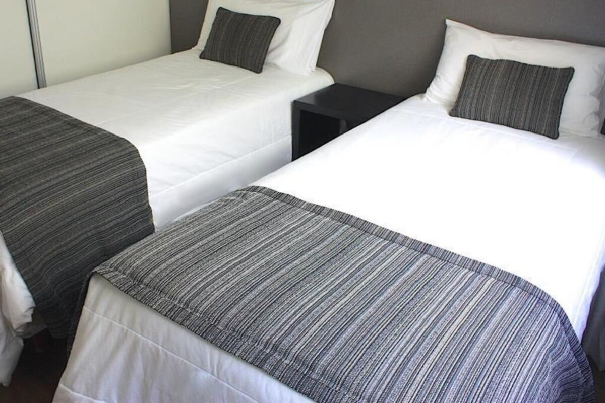 Two single beds with white linens and gray striped bedspreads, a dark nightstand, and a gray wall.