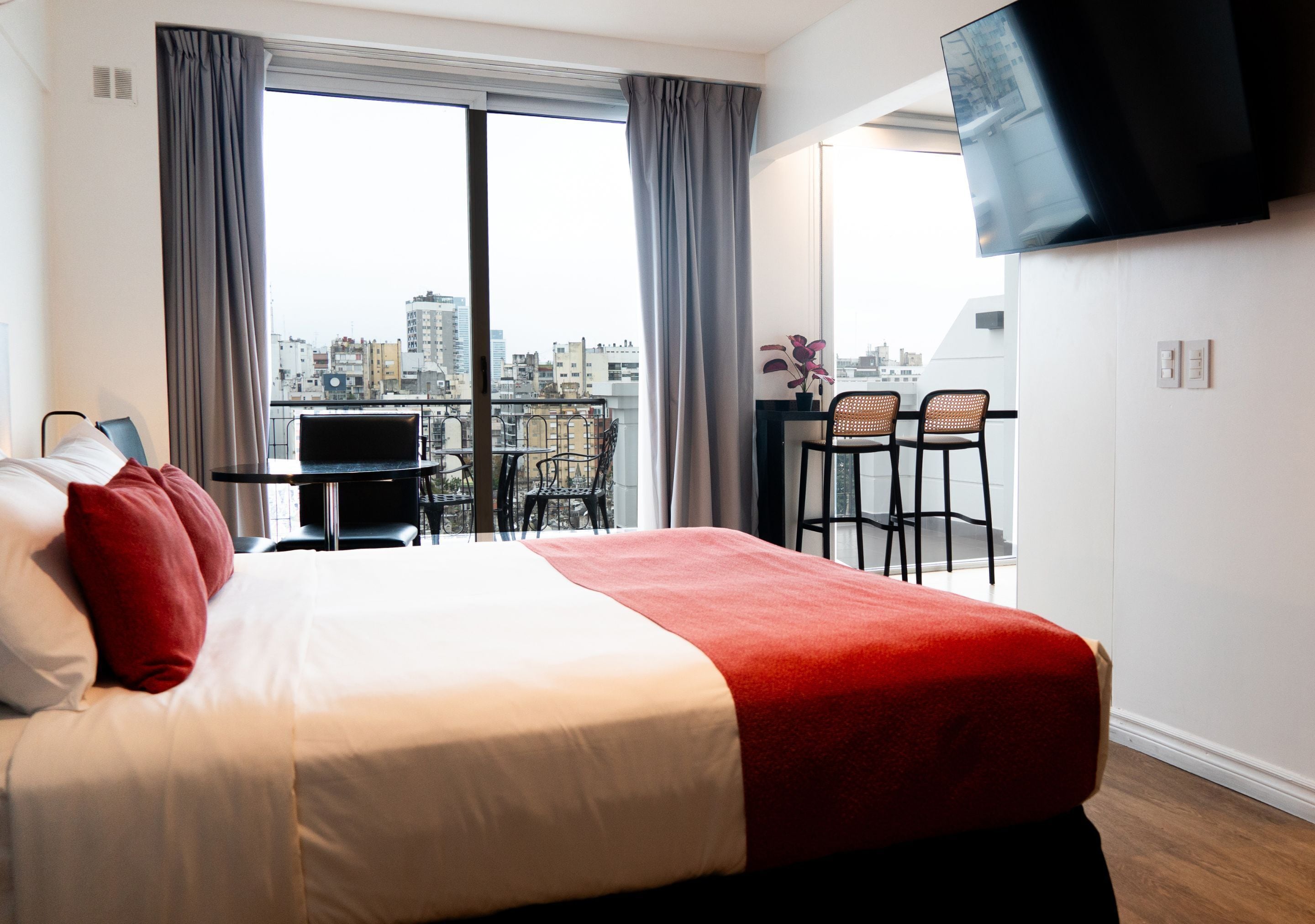 Habitación de hotel con cama, televisión, balcón con sillas y vistas a la ciudad.