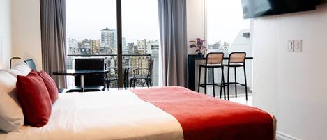 Habitación de hotel con cama, televisión, balcón con sillas y vistas a la ciudad.