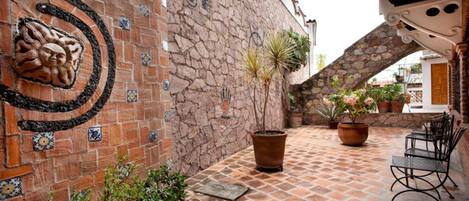 Un patio con muro de ladrillo, suelo de baldosas, plantas en macetas y mobiliario exterior.
