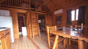 Interior de una cabaña de madera con cocina, comedor y habitación en entrepiso.
