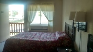 A bedroom with a bed, a nightstand, a lamp, and a view of the outdoors through a window with curtains. - Ken-Bar Lodge (Gilbertsville)