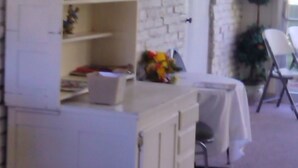 A room with a white brick wall, a white cabinet with open shelves, a small table with a white tablecloth, and a chair. - Ken-Bar Lodge (Gilbertsville)