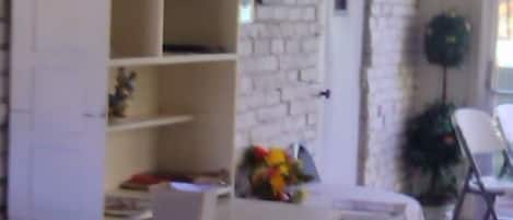 A room with a white brick wall, a white cabinet with open shelves, a small table with a white tablecloth, and a chair.