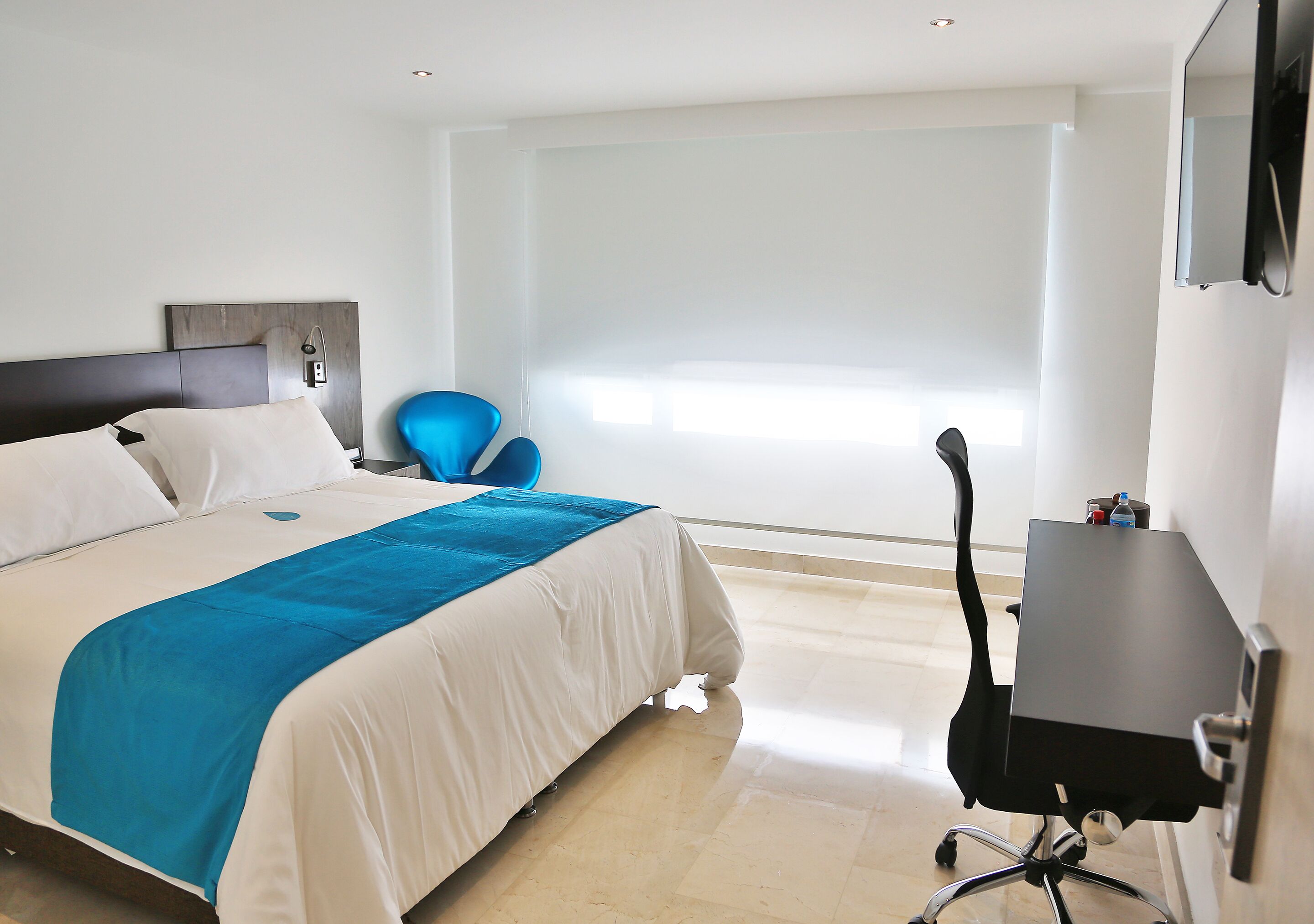A hotel room with a bed, a desk, a chair, and a large window with blinds.