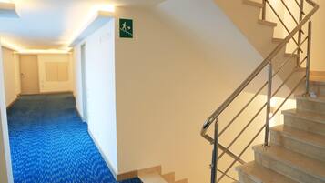 Un couloir d’hôtel avec une moquette bleue, un escalier avec des rampes en métal et un panneau indiquant la sortie de secours sur le mur.
