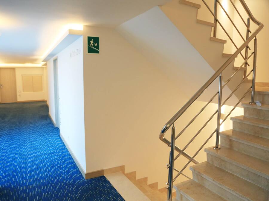 Un couloir d’hôtel avec une moquette bleue, un escalier avec des rampes en métal et un panneau indiquant la sortie de secours sur le mur.