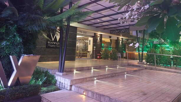 A modern hotel lobby with a glass ceiling, indoor plants, and a sign for 'Washington Plaza Hot'.