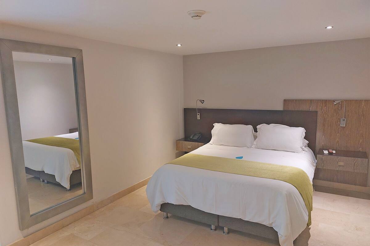 A hotel room with a large bed, a bedside table, a nightstand, a mirror, and a wooden headboard.