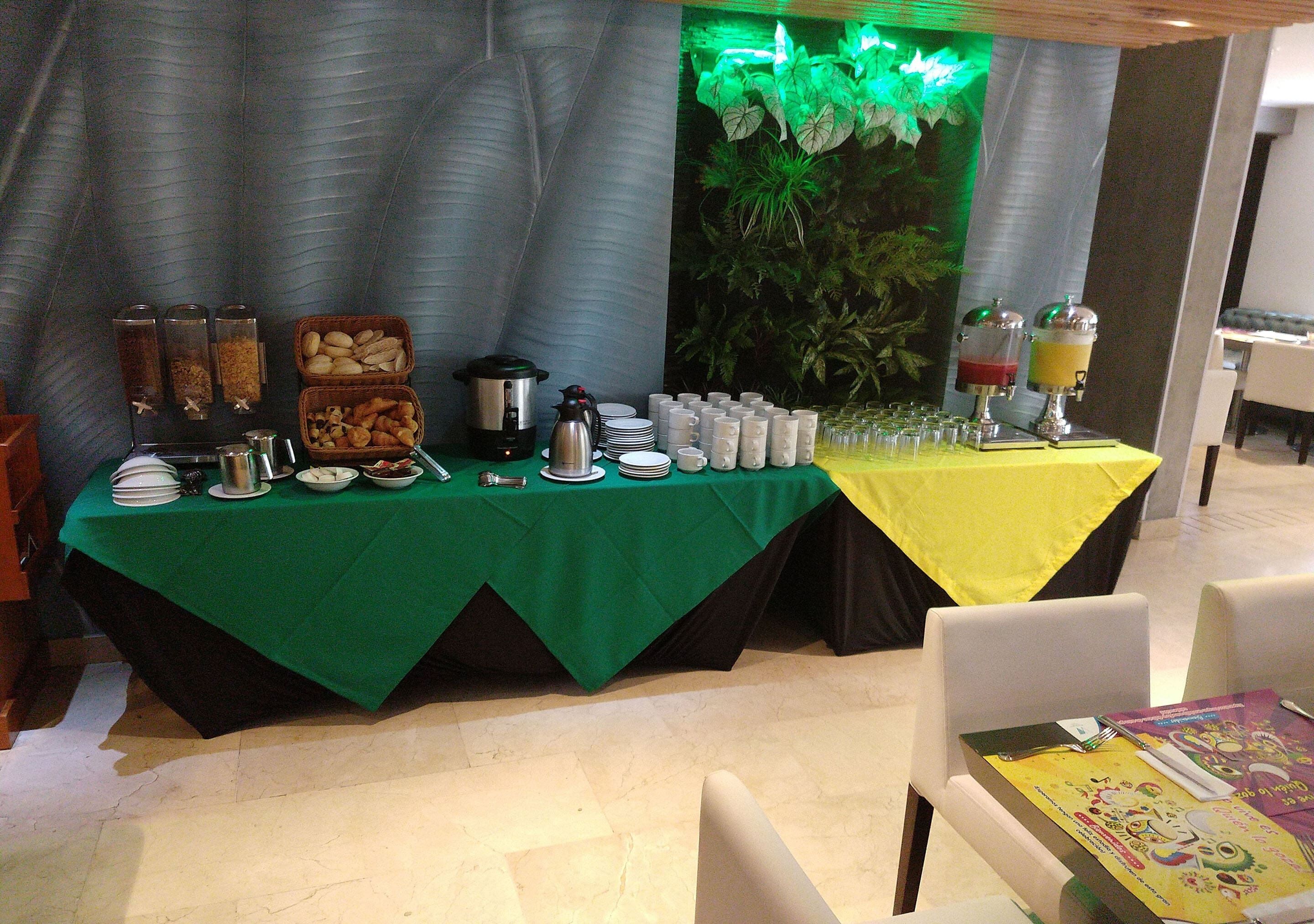 A buffet setup with various food items and beverages, a green tablecloth, and a backdrop with green foliage.