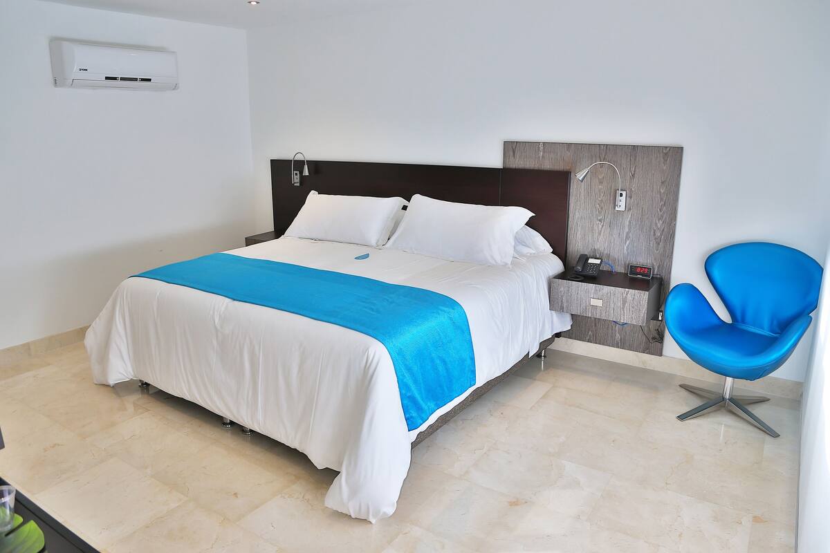 A modern hotel room with a large bed, a nightstand, a blue chair, and a wall-mounted air conditioner.