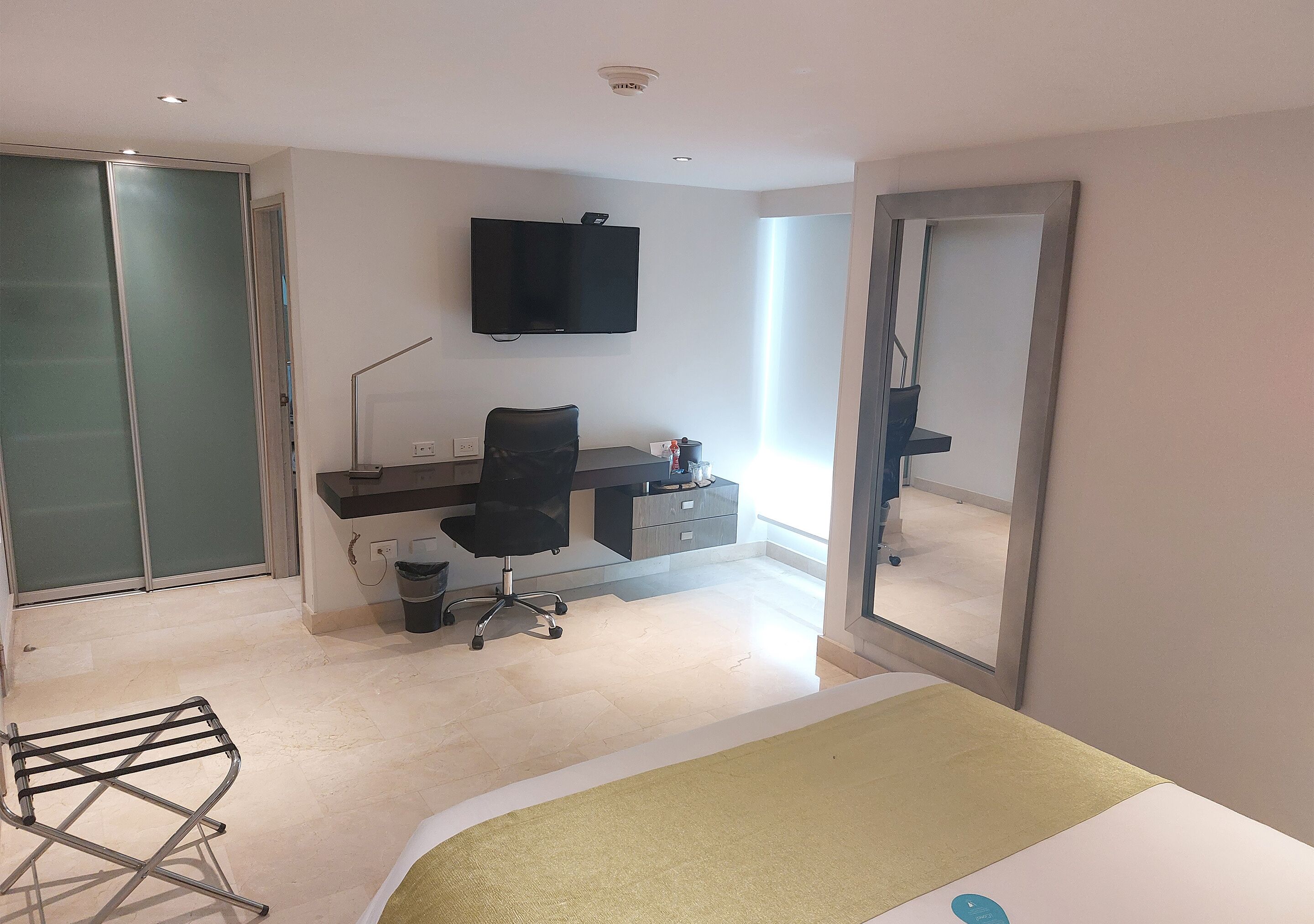 A modern hotel room with a bed, a desk with a chair, a television, and a large mirror.
