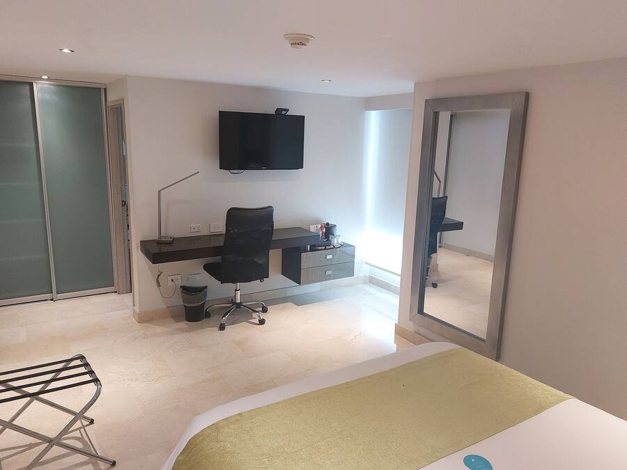 A modern hotel room with a bed, a desk with a chair, a television, and a large mirror.