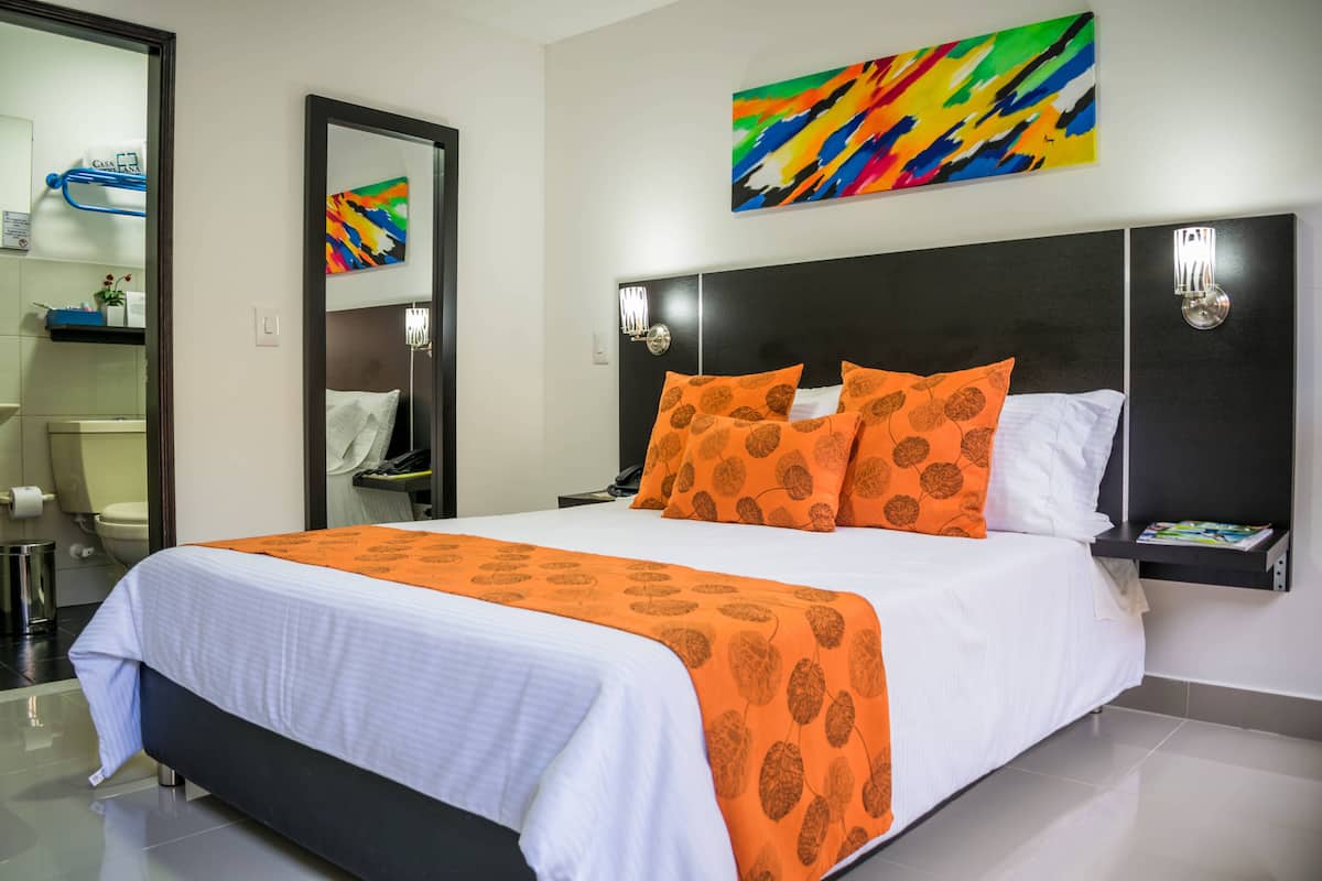 Une chambre d’hôtel moderne avec un grand lit, une salle de bain avec des toilettes et un lavabo, et une peinture abstraite aux couleurs vives accrochée au mur.