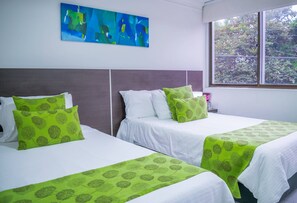 Ett hotellrum med två sängar, var och en med ett grönt, lövmönstrat överkast, vita kuddar och ett stort fönster med utsikt över grönskan utanför.
