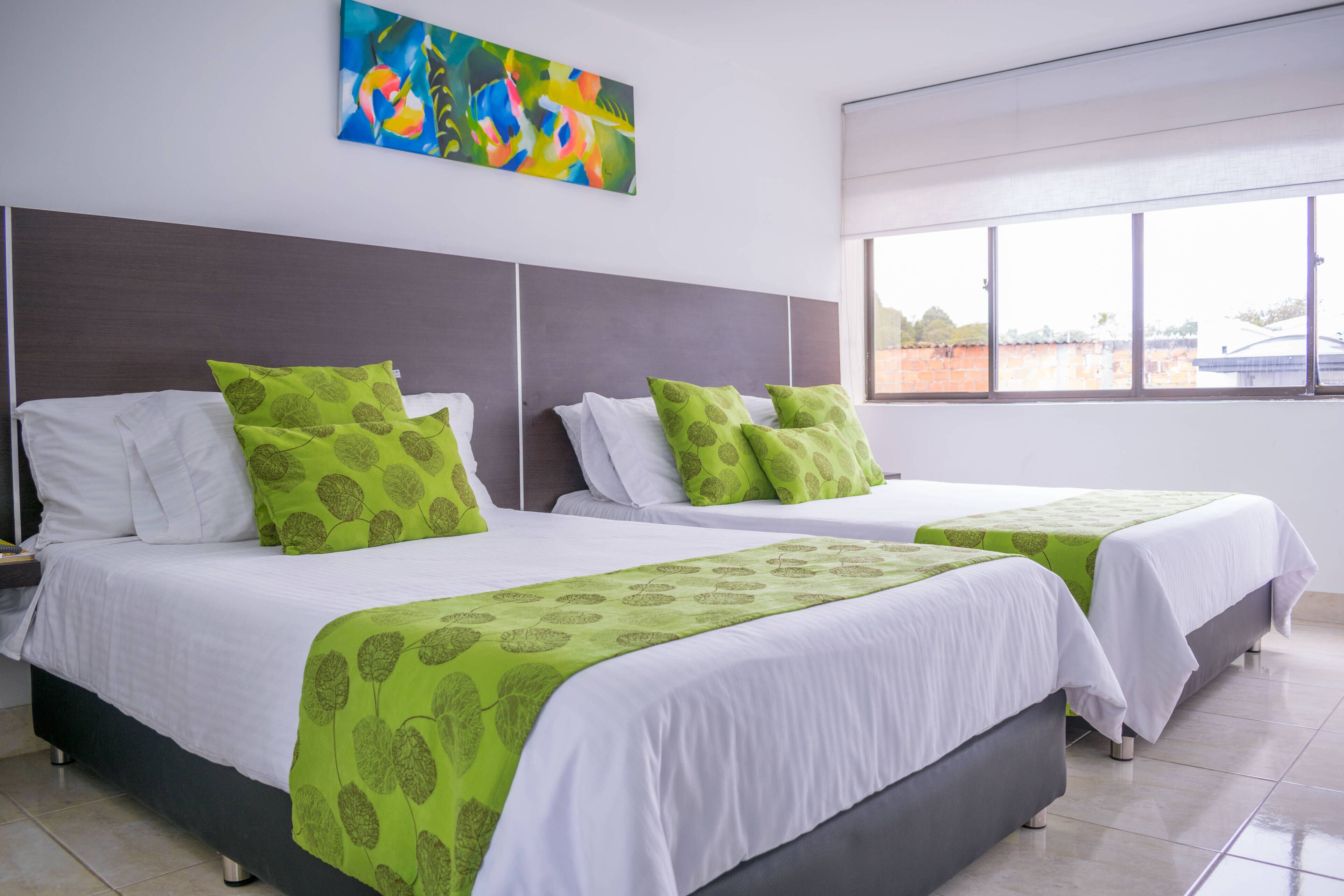 A hotel room with two beds, each with white bedding and green leaf-patterned throws. There is a large window providing a view of buildings outside.