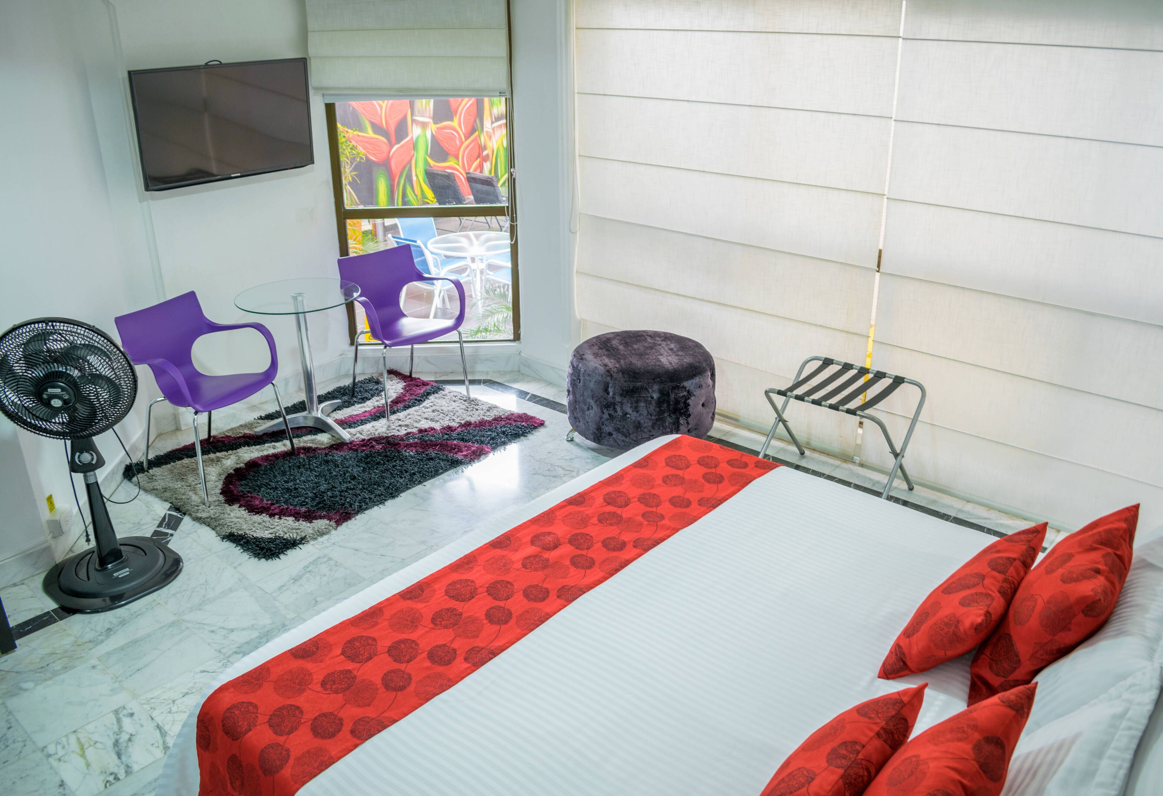 Hotellihuone, jossa on sänky, tuuletin, violetti tuoli, pieni pöytä ja ikkuna, josta avautuu näkymä värikkäisiin kukkiin.