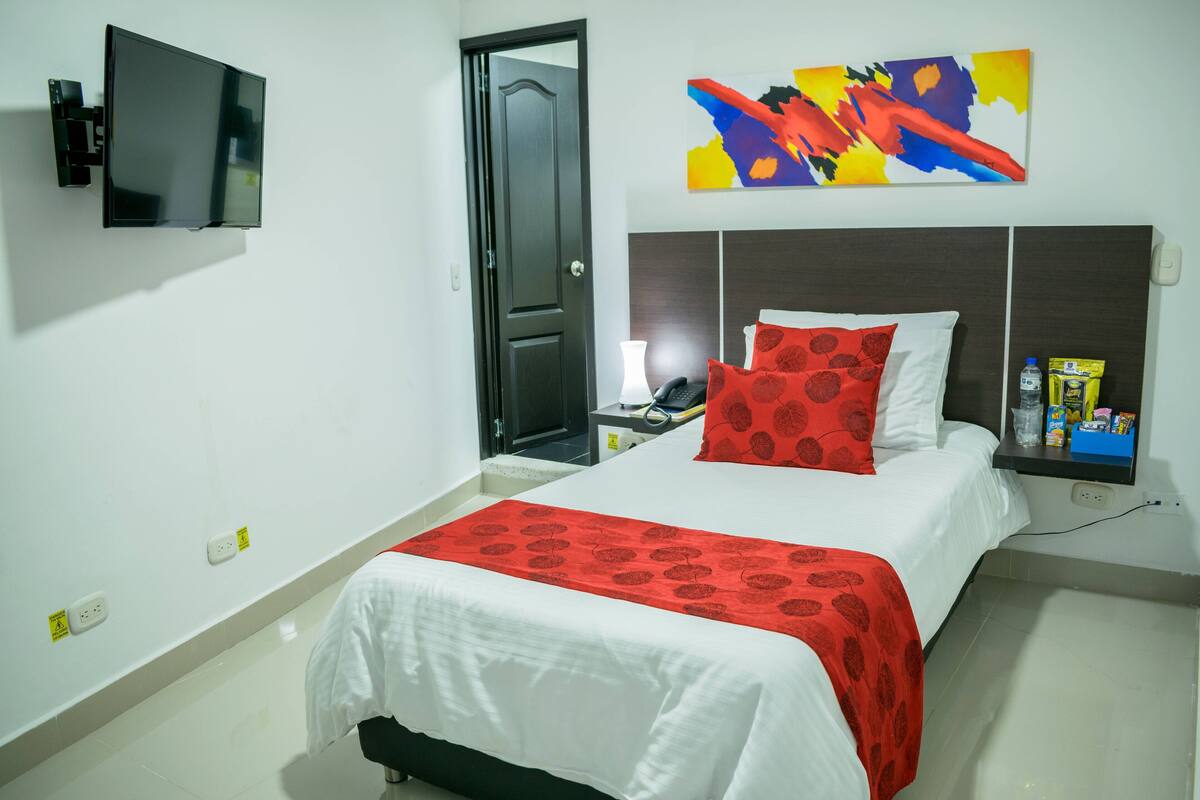 Une chambre d’hôtel avec un lit, un téléviseur fixé au mur, une porte, un tableau et une étagère avec quelques objets.
