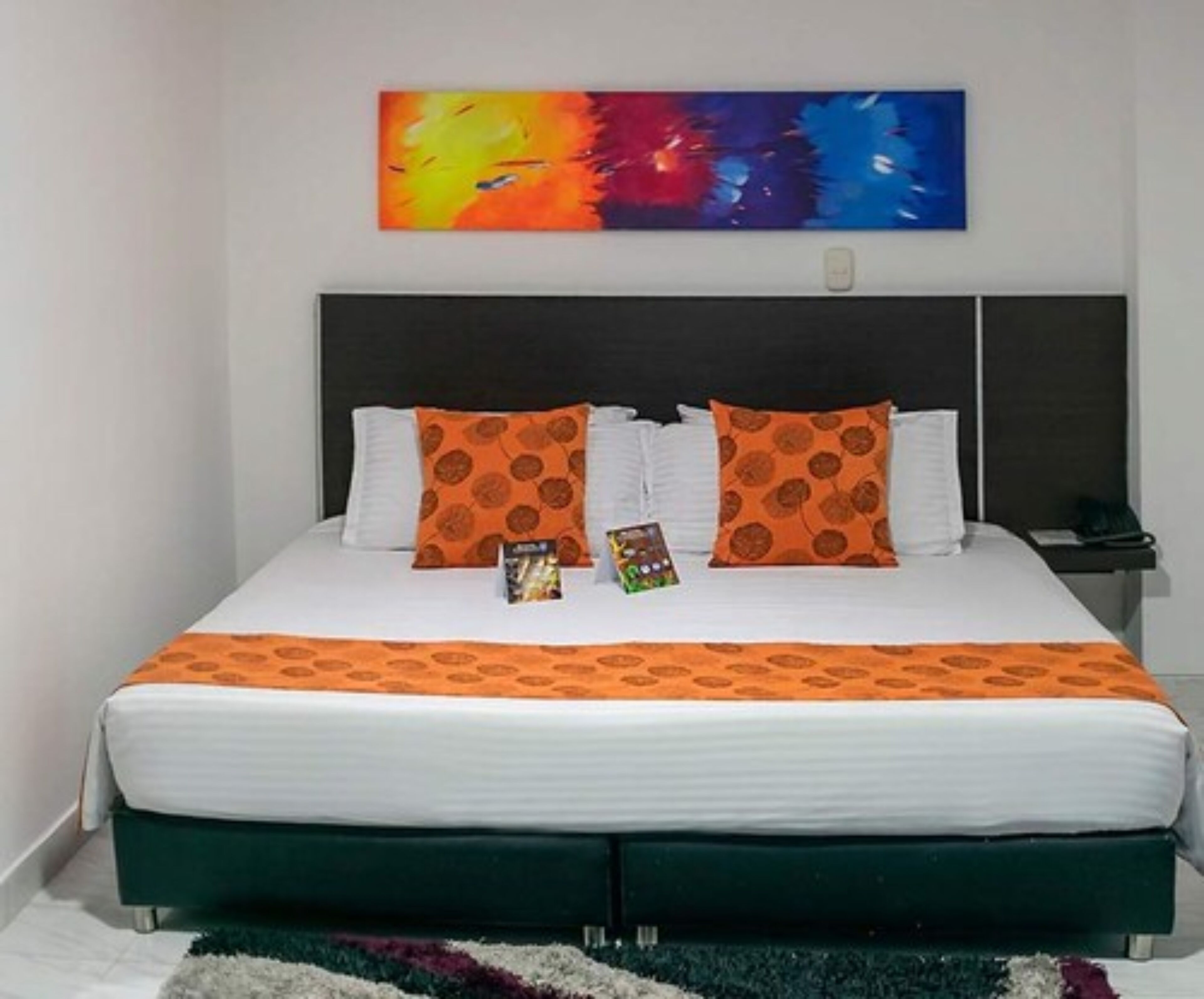 Una cama con colcha naranja estampada, dos almohadas a juego y un cuadro abstracto colorido en la pared de arriba.
