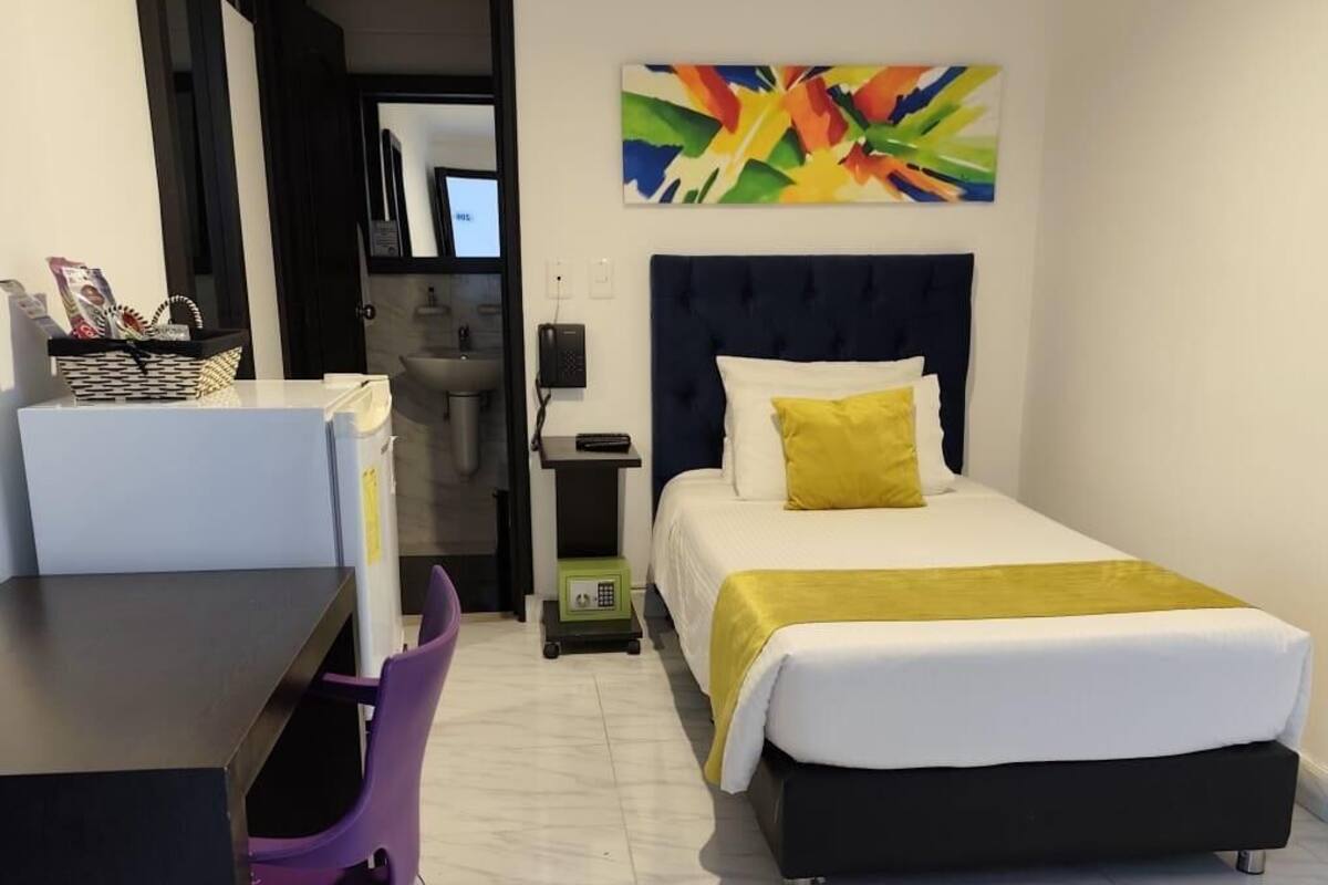 Une chambre d’hôtel avec un lit, un bureau, une chaise, une petite table, un tableau et une salle de bain visible à travers une porte ouverte.