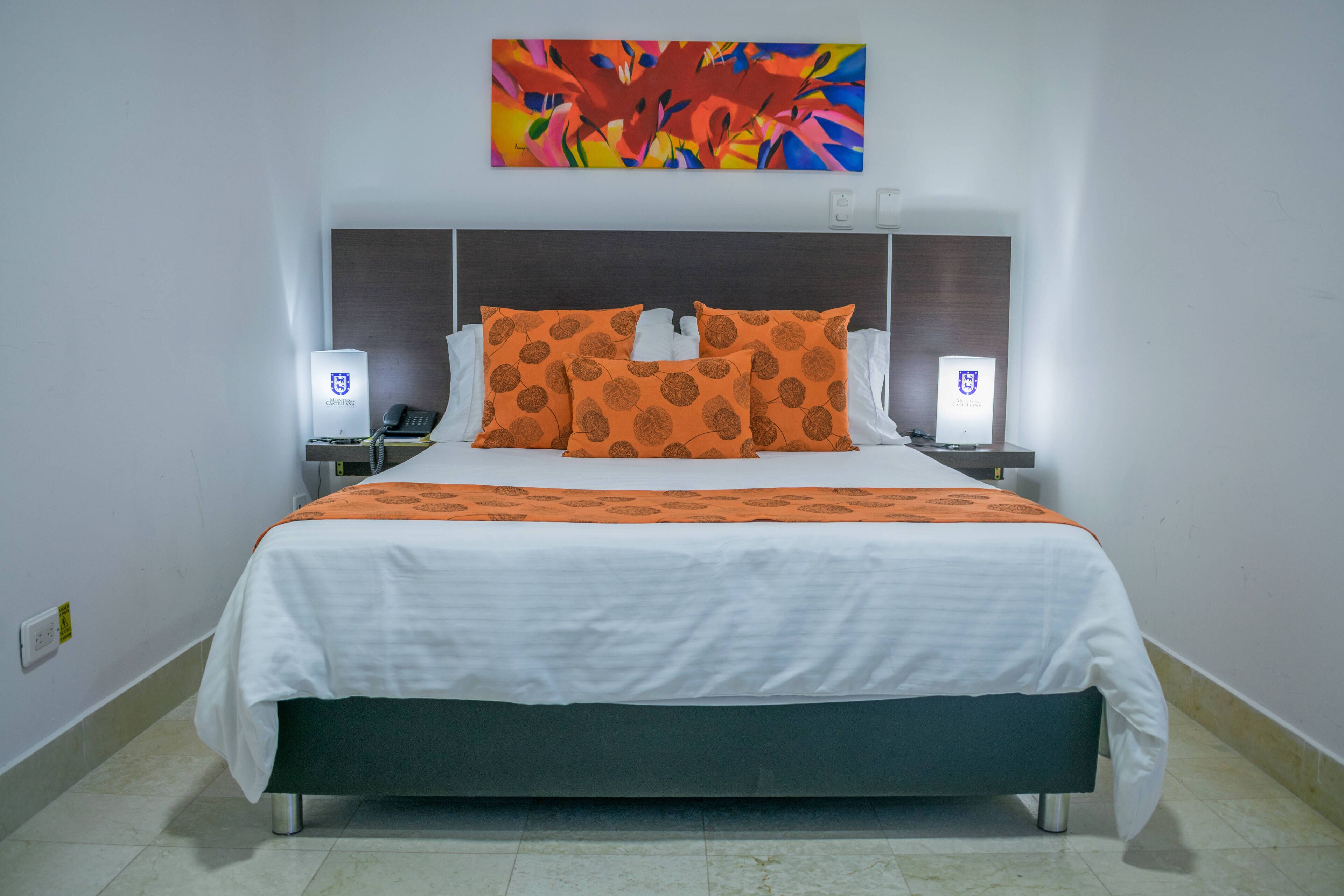 Habitación de hotel con una cama grande, dos mesitas de noche, un cuadro abstracto de colores sobre la cama y una pared blanca.