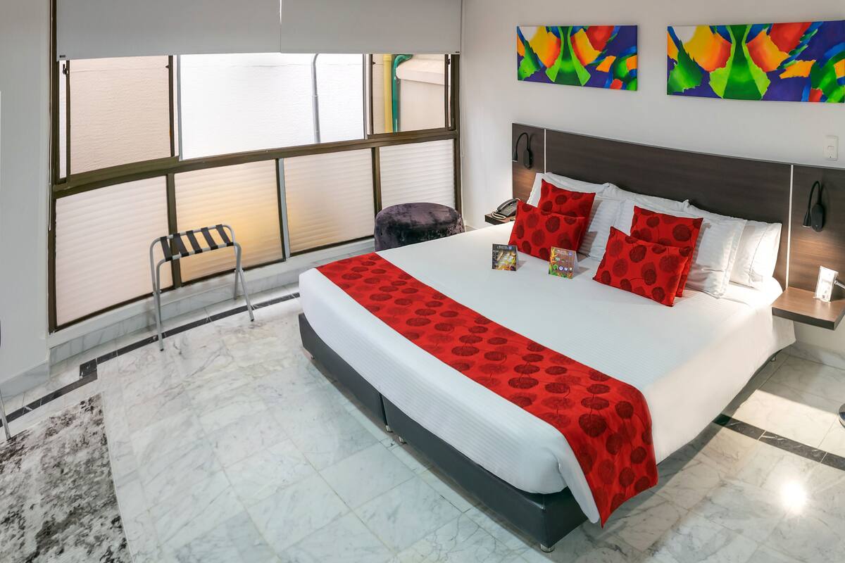 Une chambre d’hôtel moderne avec un grand lit, une literie rouge et blanche, une vue par une fenêtre avec des stores, et une œuvre d’art abstraite au mur.
