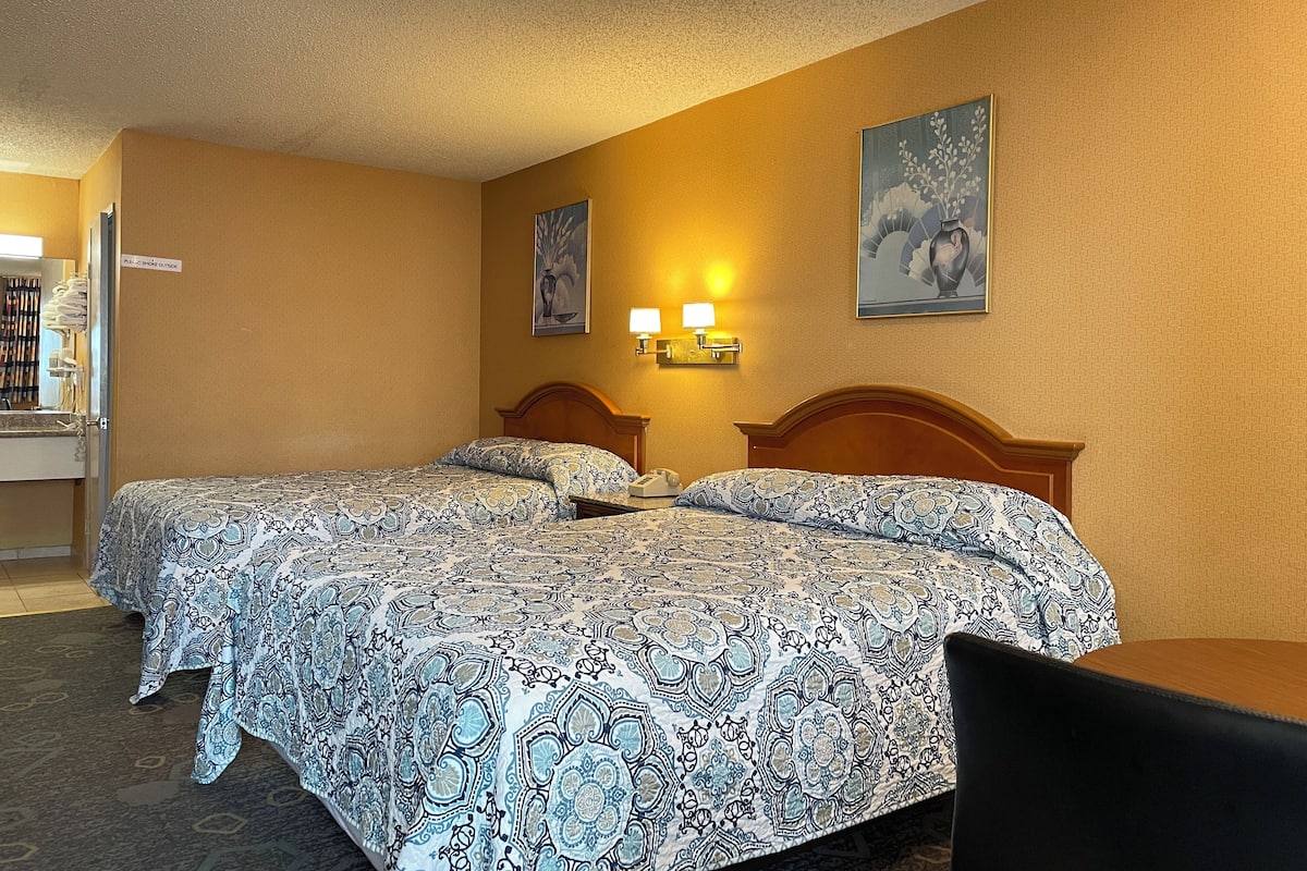 A hotel room with two beds, a desk, and a chair. There are two wall-mounted lamps and a painting above the beds.
