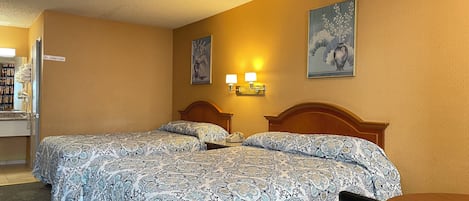 A hotel room with two beds, a desk, and a chair. There are two wall-mounted lamps and a painting above the beds.