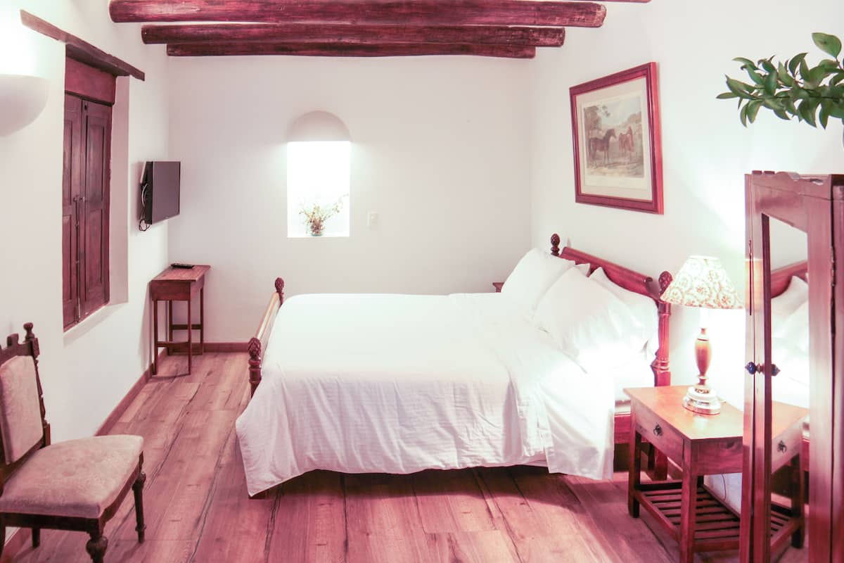 Un dormitorio con una cama de madera, una silla, una mesita de noche con una lámpara, una pequeña mesa con un florero y un cuadro en la pared.