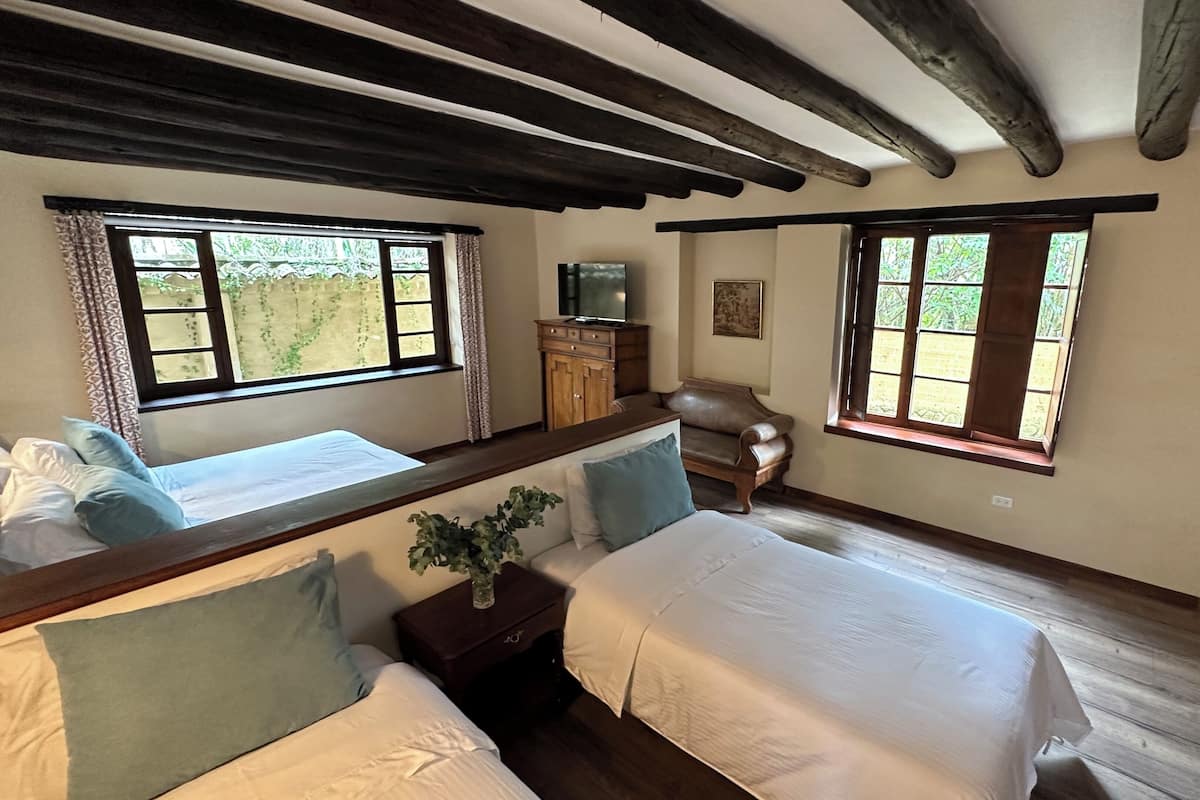 Habitación con dos camas, vigas de madera en el techo, un escritorio con silla y ventanas que dan a zonas verdes.