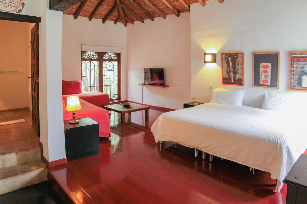 Una habitación con una cama, un sofá rojo, una mesita con una lámpara y un televisor sobre una estantería.