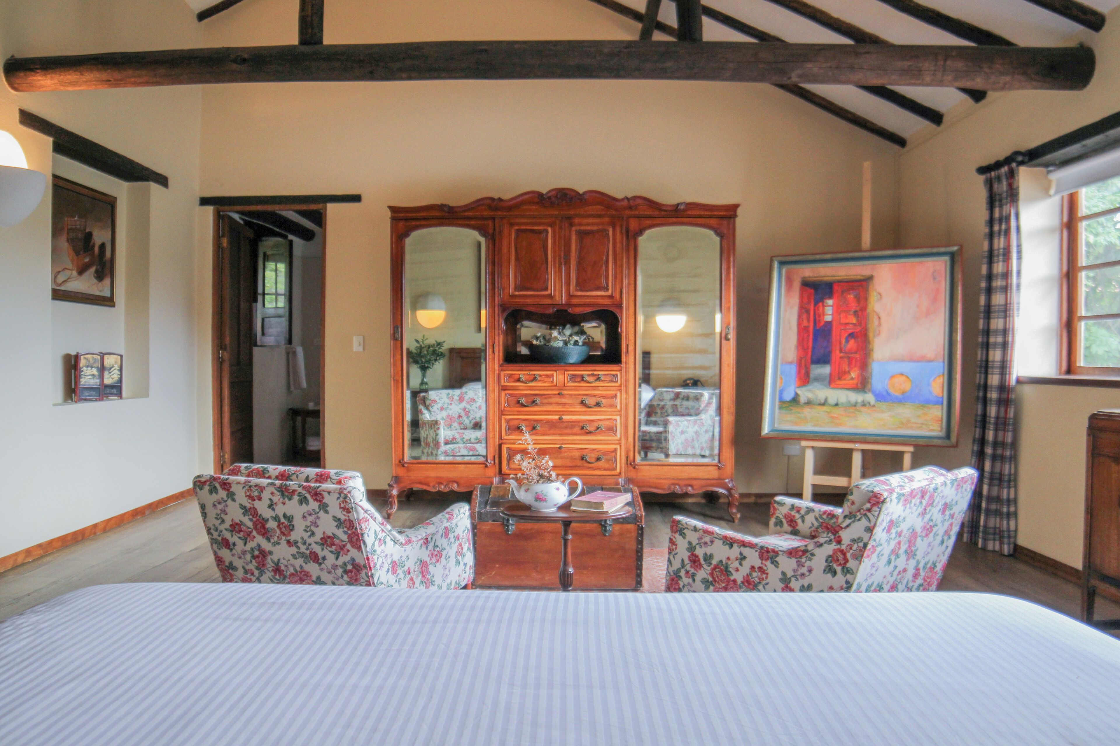 A bedroom with a large bed, floral-patterned chairs, a wooden dresser with mirrors, and a painting on the wall.