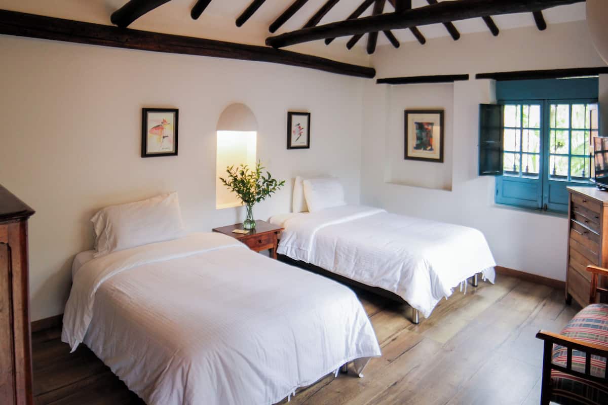 Una habitación con dos camas, pisos de madera, un escritorio con silla y una ventana con contraventanas azules.