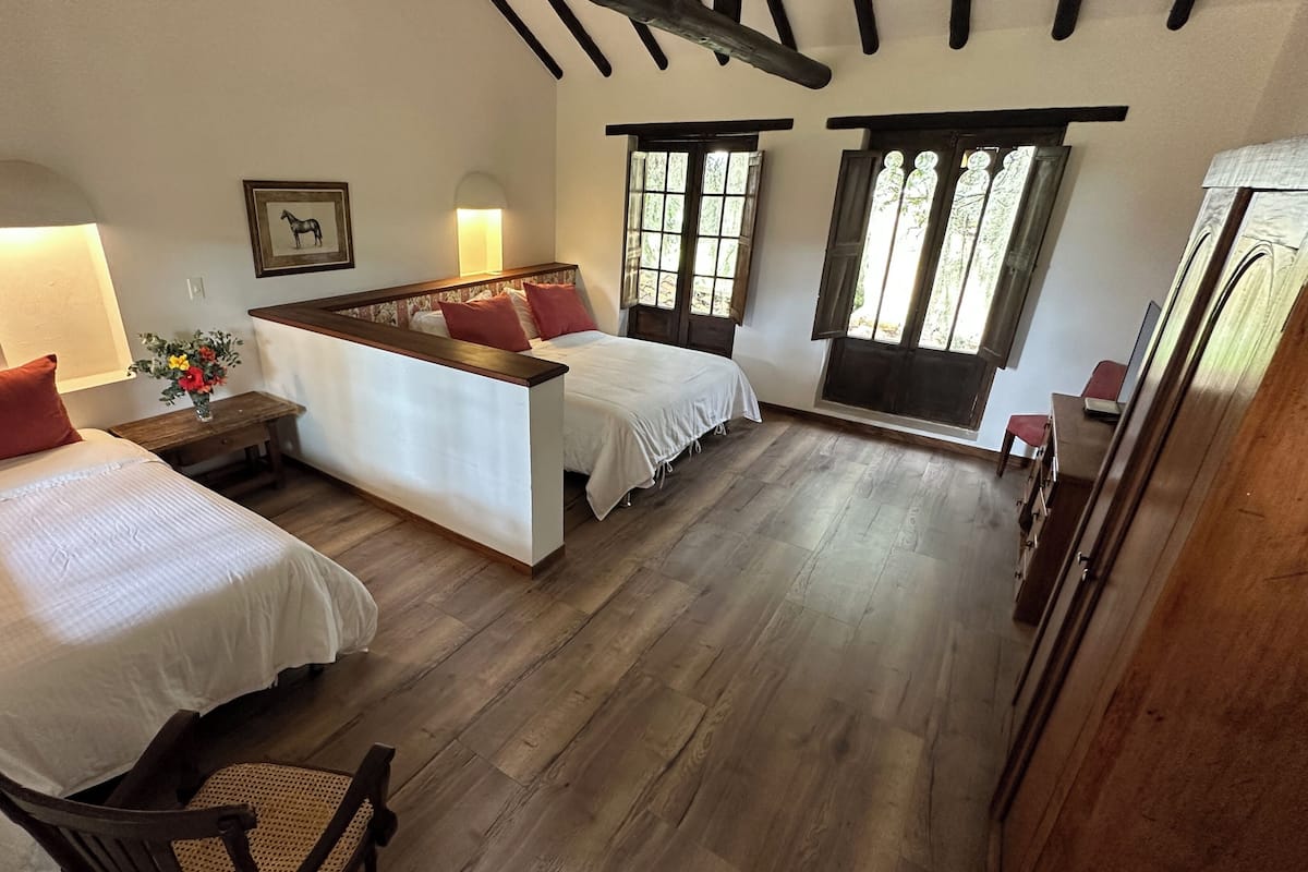 Una habitación con dos camas, un banco de madera, una mesita con un florero y un ventanal con vistas al verdor.