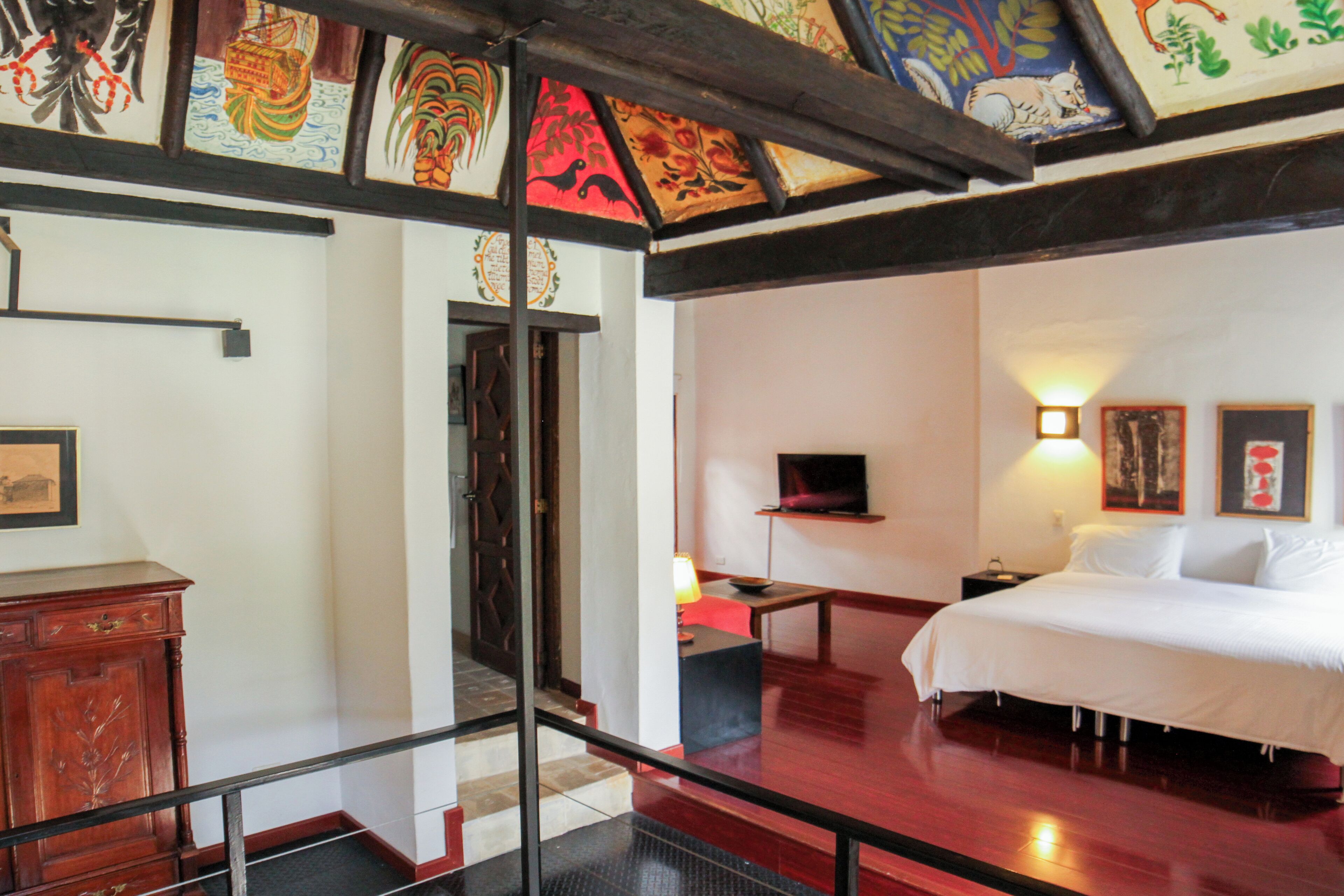 Ein Hotelzimmer mit Bett, Fernseher, Schreibtisch und Stuhl. Es gibt einen Holzkasten und eine Tür mit einer Zierausstattung.