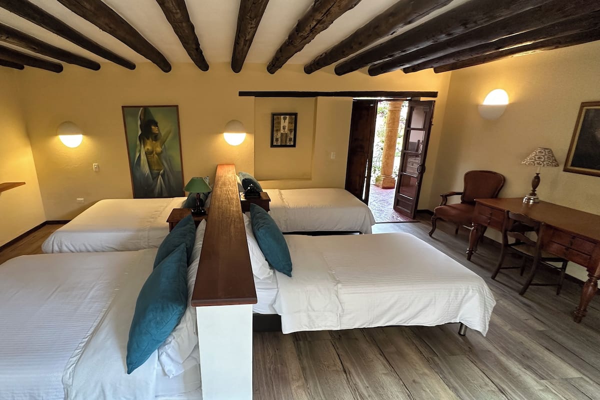 Habitación de hotel con dos camas, una mesa de madera, un escritorio, una silla, un televisor y obras de arte en las paredes.