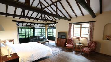 A bedroom with a large bed, a TV, a wooden ceiling, and a view of greenery through the windows.