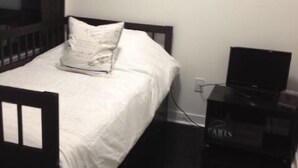 Ein Einzelbett mit Holzrahmen und weißer Bettwäsche, ein schwarzer TV-Schrank mit Flachbildfernseher und eine schlichte Wand mit Steckdose.