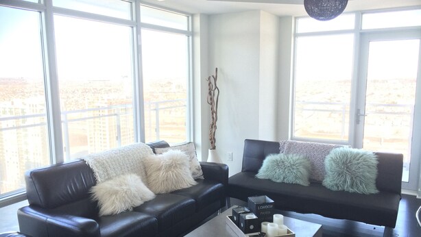 Ein modernes Wohnzimmer mit zwei schwarzen Ledersofas, weißen, flauschigen Kissen und einem Couchtisch mit Kerzen. Große Fenster bieten Blick auf eine Stadtlandschaft.