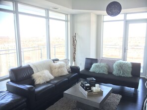 Un salon moderne avec deux canapés en cuir noir, des coussins blancs et moelleux, et une table basse agrémentée de bougies. De grandes fenêtres offrent une vue sur le paysage urbain.