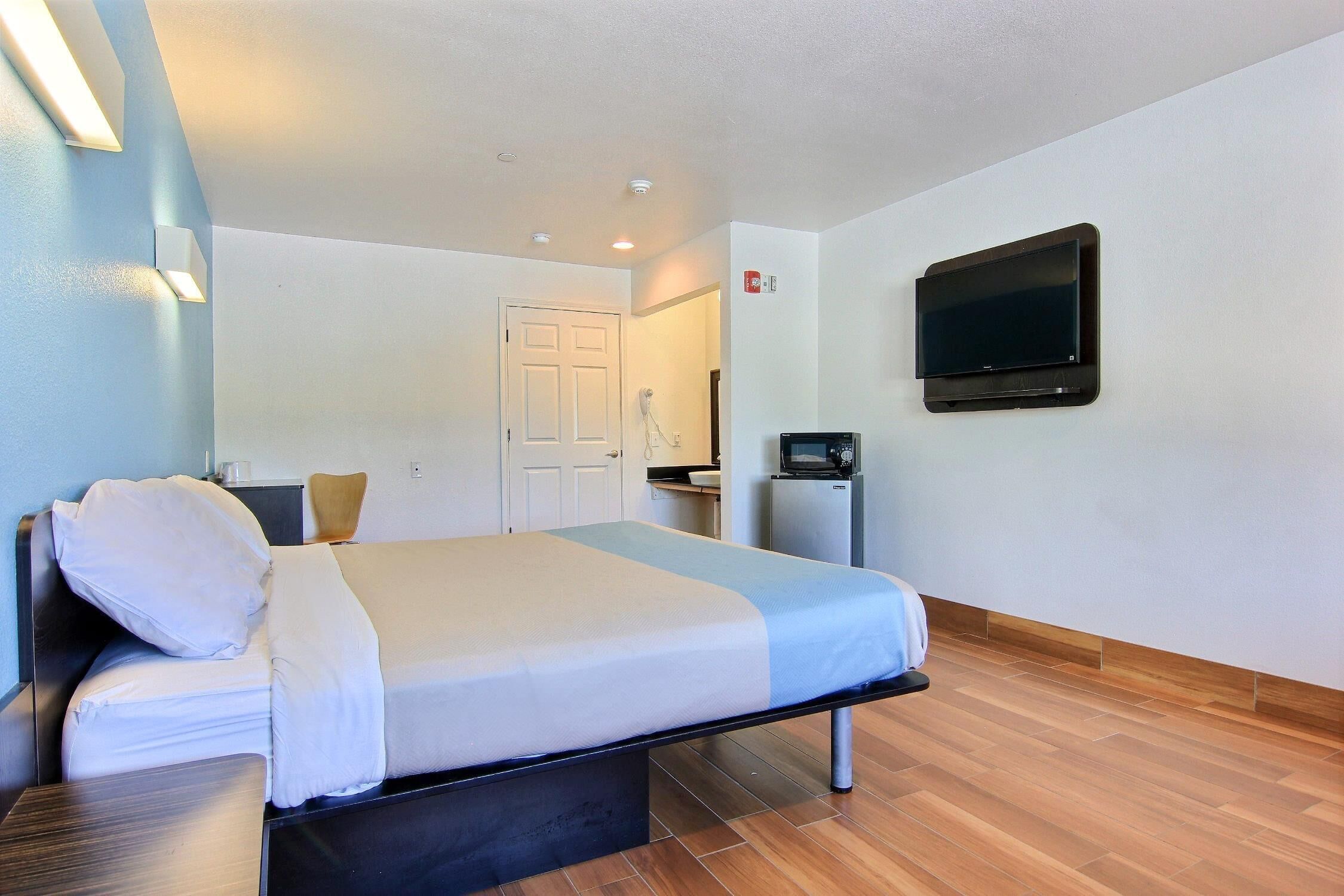 A hotel room with a bed, a flat-screen TV mounted on the wall, a small refrigerator, and a door leading to a bathroom.