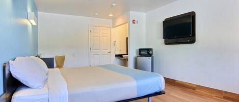 A hotel room with a bed, a flat-screen TV mounted on the wall, a small refrigerator, and a door leading to a bathroom.