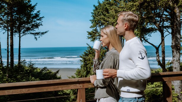 A couple standing on a wooden deck overlooking a beach with ocean waves and trees in the background. - Ocean Crest Resort (Moclips)