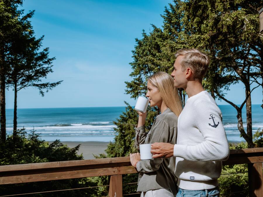 Ein Paar steht auf einer Holzterrasse mit Blick auf einen Strand, wo Meereswellen und Bäume im Hintergrund zu sehen sind.