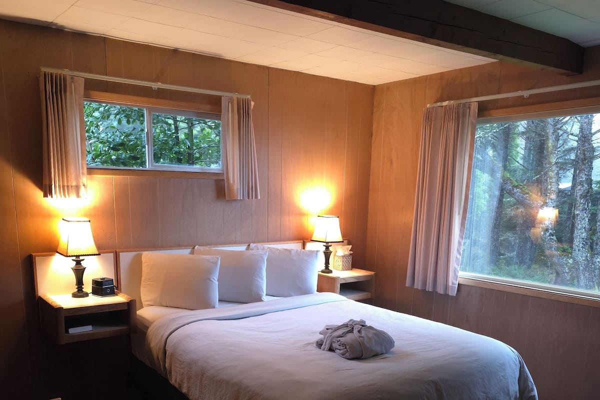 A bedroom with a bed, two bedside tables with lamps, a window with curtains, and a view of trees.