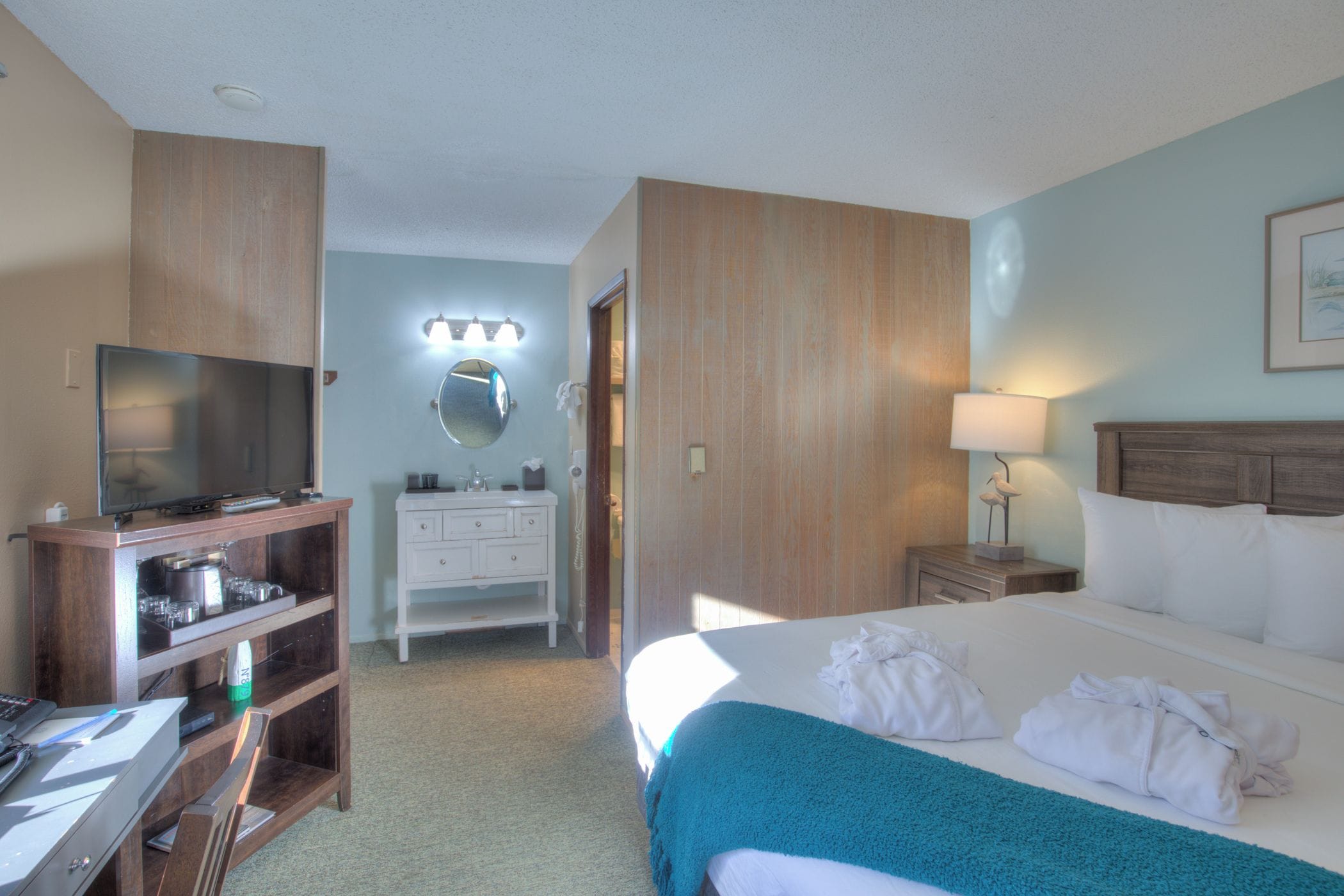 A hotel room with a bed, a desk with a computer, a television, a vanity with a mirror, and a bedside lamp.