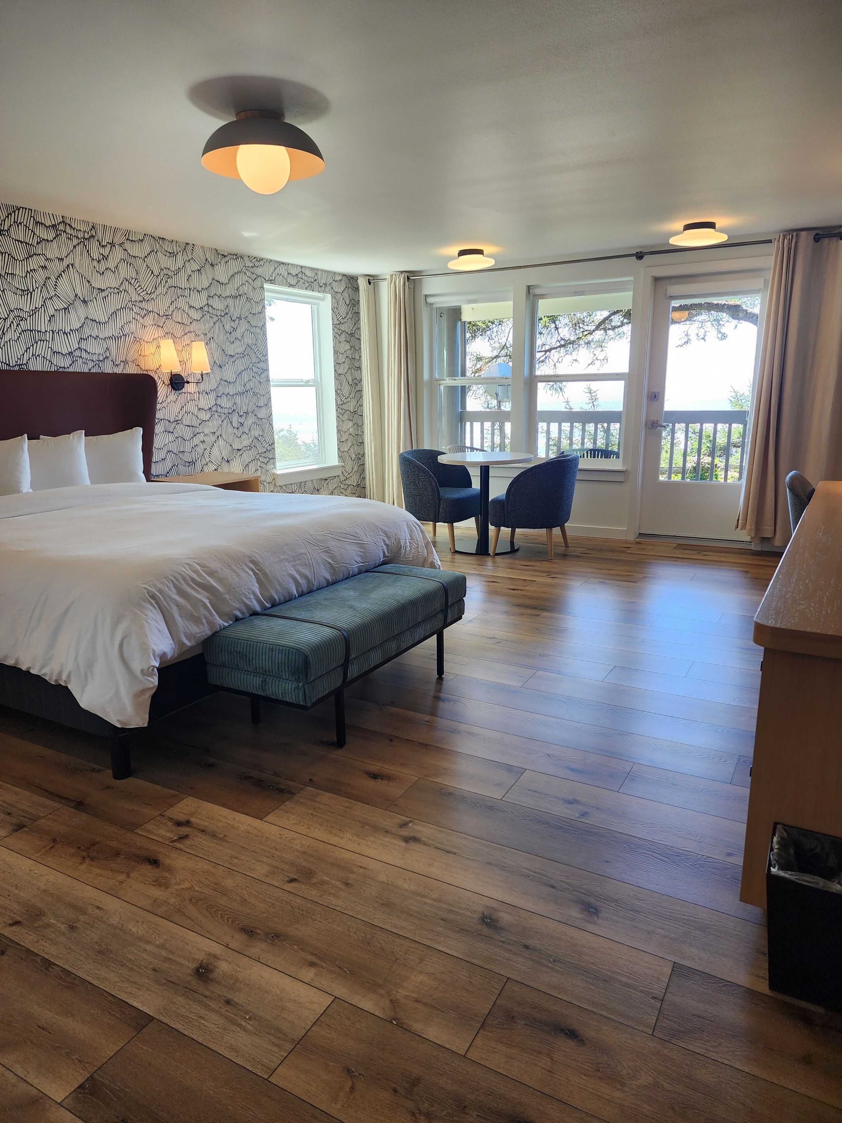 A hotel room with a large bed, a bench, two chairs, a dining table, and a view through the glass doors.